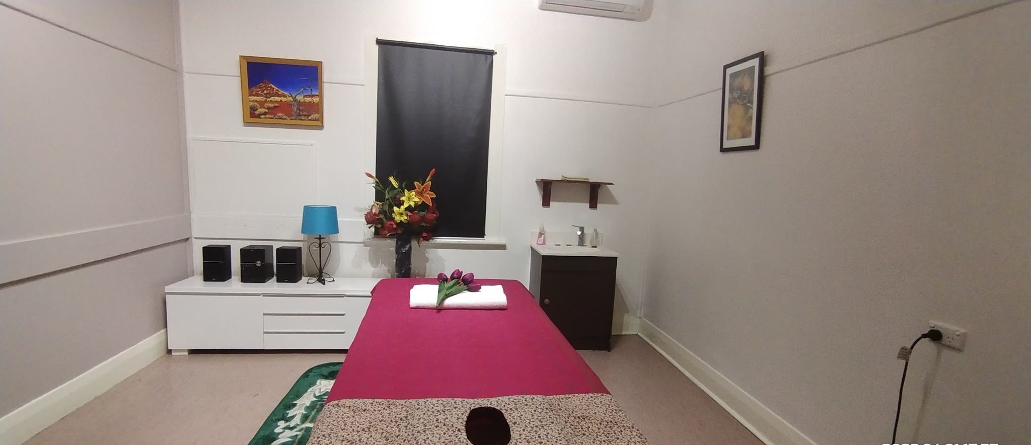 A second treatment room with a massage table dressed in deep pink, a vase of lilies and a desert painting