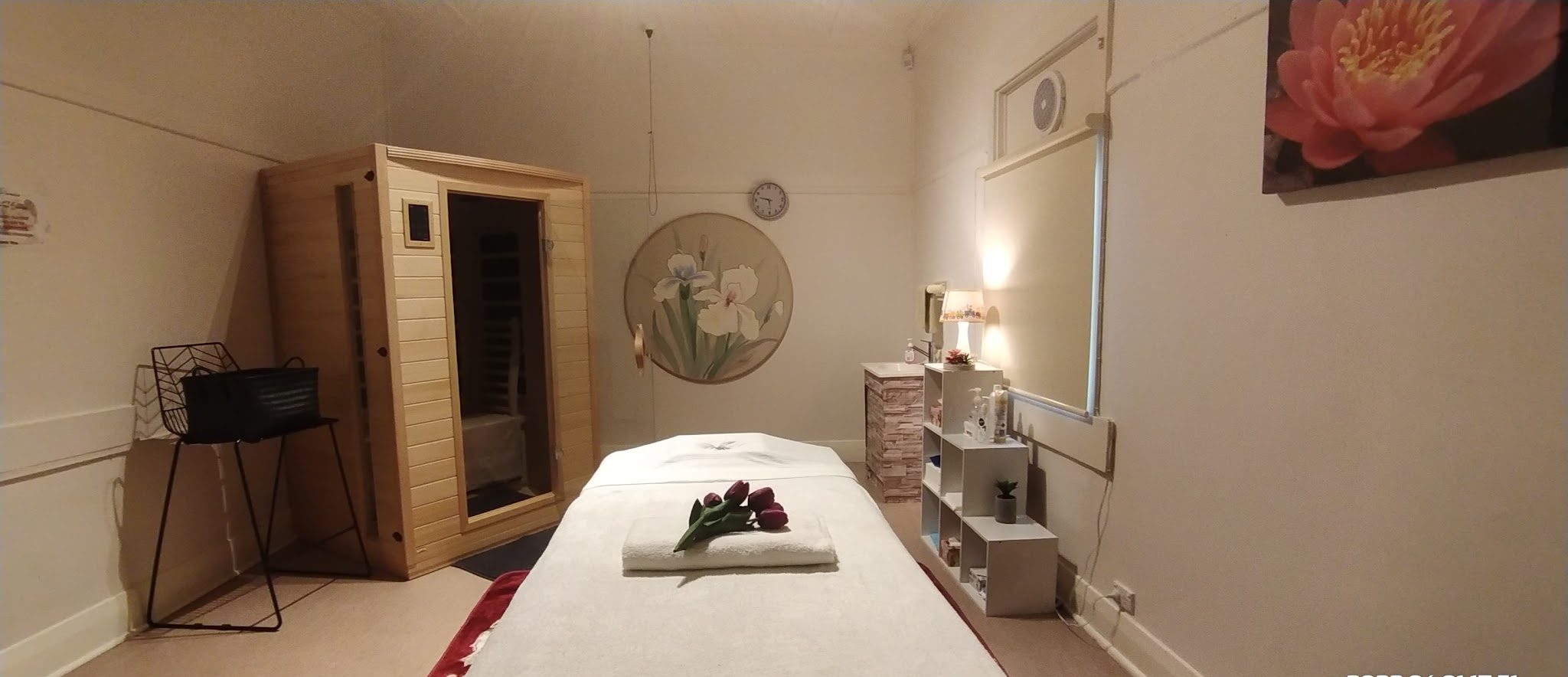 A treatment room with a massage table, folded towel and tulips, and the infrared sauna cabin in the corner